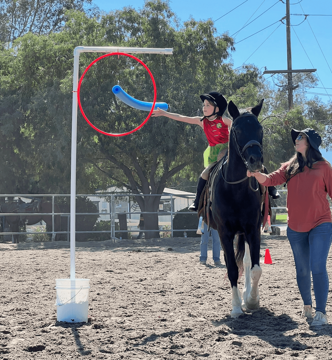 A small rider on a black horse reaching to push a pool noodle through a red hoop while a handler leads