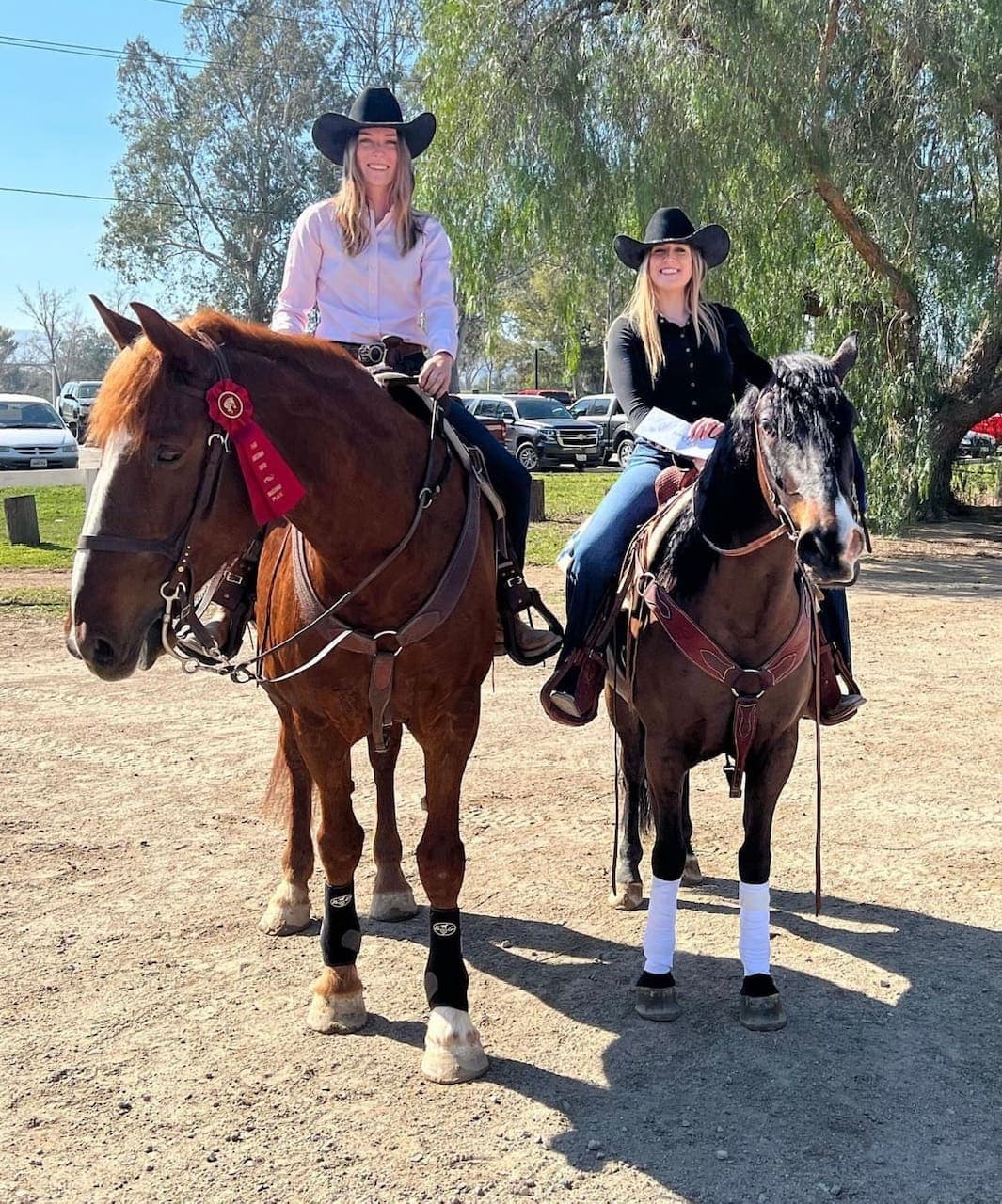 Two riders in western hats on their horses, one wearing a red show ribbon