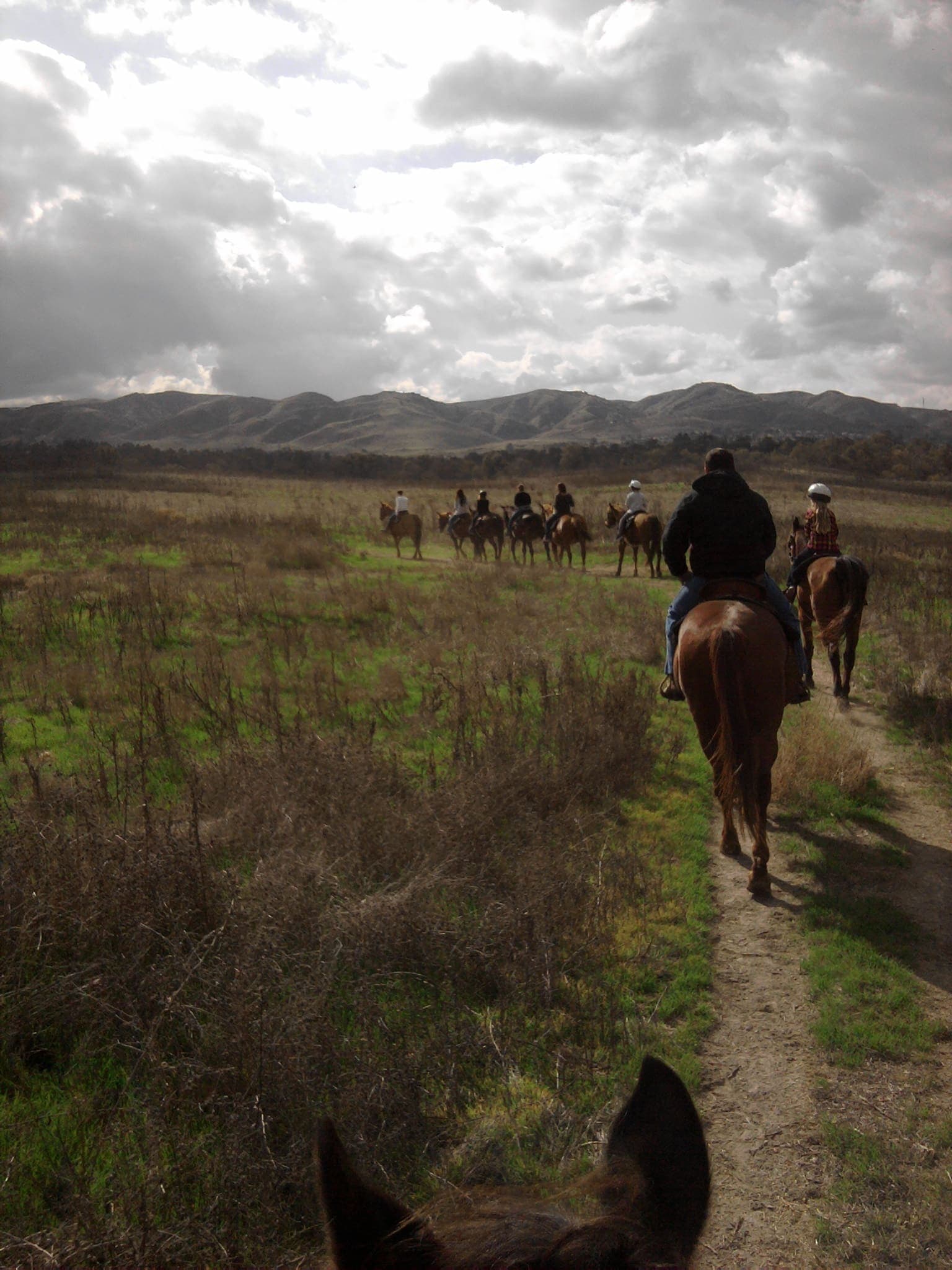 A line of riders heading up the trail toward the hills under dramatic winter clouds, photographed between a horse’s ears